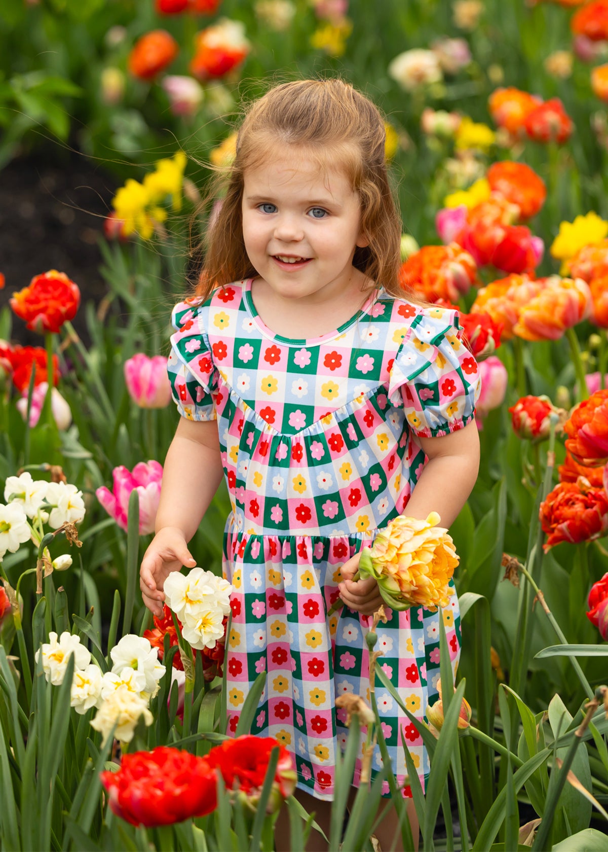 A young girl wears the vibrant Flower Pops Short Sleeve Dress by Mabel and Honey, standing among red, orange, yellow, and white flowers while holding a bouquet and smiling at the camera on a bright day.