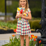 A young girl wears the Mabel and Honey Flower Pops Cap Sleeve Two Piece Set, smiling among blooming tulips with a bunch of yellow tulips in hand. She pairs it with white socks and shoes, standing in front of a garden and black fence.