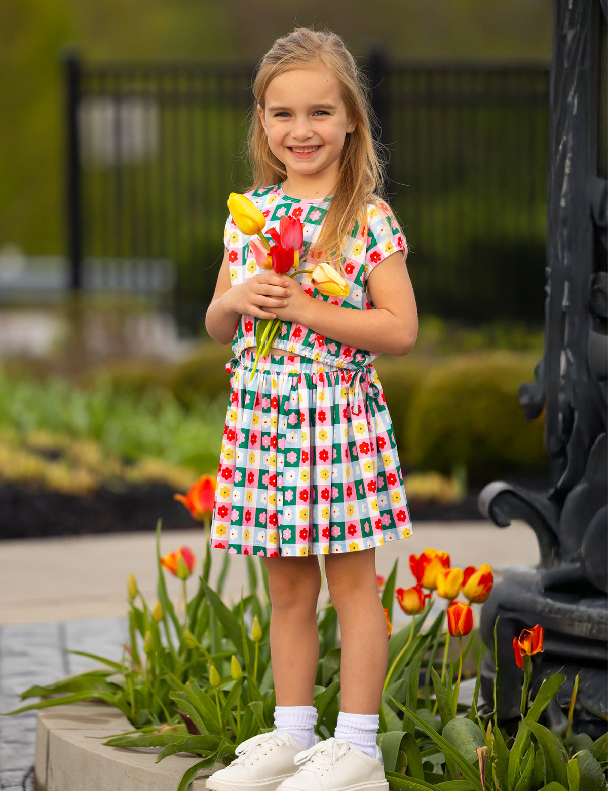 A young girl wears the Mabel and Honey Flower Pops Cap Sleeve Two Piece Set, smiling among blooming tulips with a bunch of yellow tulips in hand. She pairs it with white socks and shoes, standing in front of a garden and black fence.