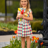 A young girl wears the Mabel and Honey Flower Pops Cap Sleeve Two Piece Set, smiling among blooming tulips with a bunch of yellow tulips in hand. She pairs it with white socks and shoes, standing in front of a garden and black fence.