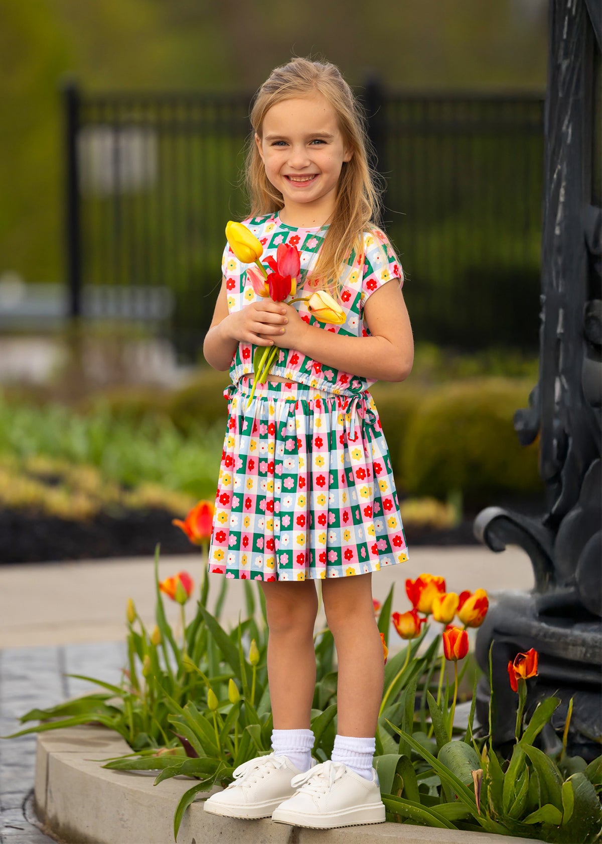 A young girl wears the Mabel and Honey Flower Pops Cap Sleeve Two Piece Set, smiling among blooming tulips with a bunch of yellow tulips in hand. She pairs it with white socks and shoes, standing in front of a garden and black fence.