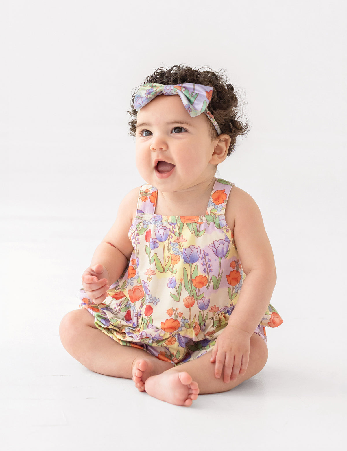 A smiling baby with curly dark hair wears a floral headband and the Mabel and Honey Tulip Twirl Tank Romper, featuring a colorful knit design and tulip waist, while sitting on the floor against a plain white background.