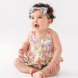 A smiling baby with curly dark hair wears a floral headband and the Mabel and Honey Tulip Twirl Tank Romper, featuring a colorful knit design and tulip waist, while sitting on the floor against a plain white background.