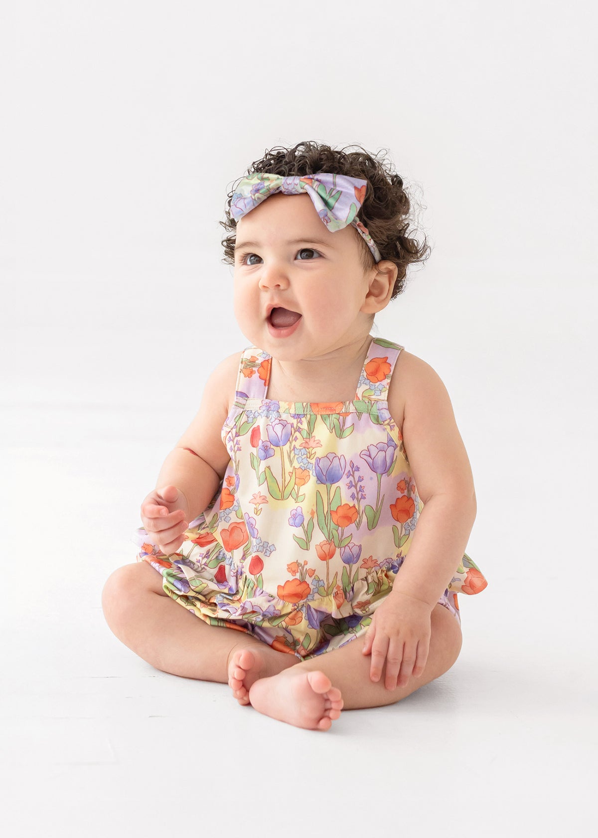 A smiling baby with curly dark hair wears a floral headband and the Mabel and Honey Tulip Twirl Tank Romper, featuring a colorful knit design and tulip waist, while sitting on the floor against a plain white background.