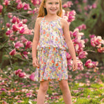 A young girl smiles among blooming pink magnolias, wearing the vibrant Tulip Twirl Sleeveless Dress by Mabel and Honey, with green grass and scattered petals at her feet.