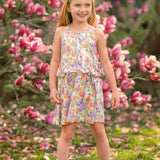 A young girl smiles among blooming pink magnolias, wearing the vibrant Tulip Twirl Sleeveless Dress by Mabel and Honey, with green grass and scattered petals at her feet.