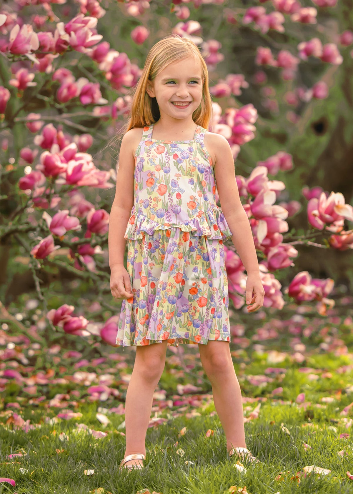 A young girl smiles among blooming pink magnolias, wearing the vibrant Tulip Twirl Sleeveless Dress by Mabel and Honey, with green grass and scattered petals at her feet.