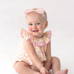A smiling baby sits on the floor wearing Mabel and Honey’s Ashley Cap Sleeve Romper in pink and yellow plaid with ruffled sleeves and a matching headband. The plain white background highlights this adorable 100% cotton outfit.