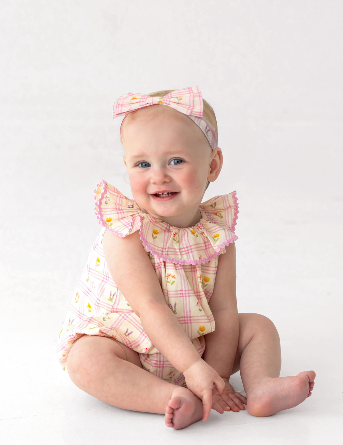 A smiling baby sits on the floor wearing Mabel and Honey’s Ashley Cap Sleeve Romper in pink and yellow plaid with ruffled sleeves and a matching headband. The plain white background highlights this adorable 100% cotton outfit.