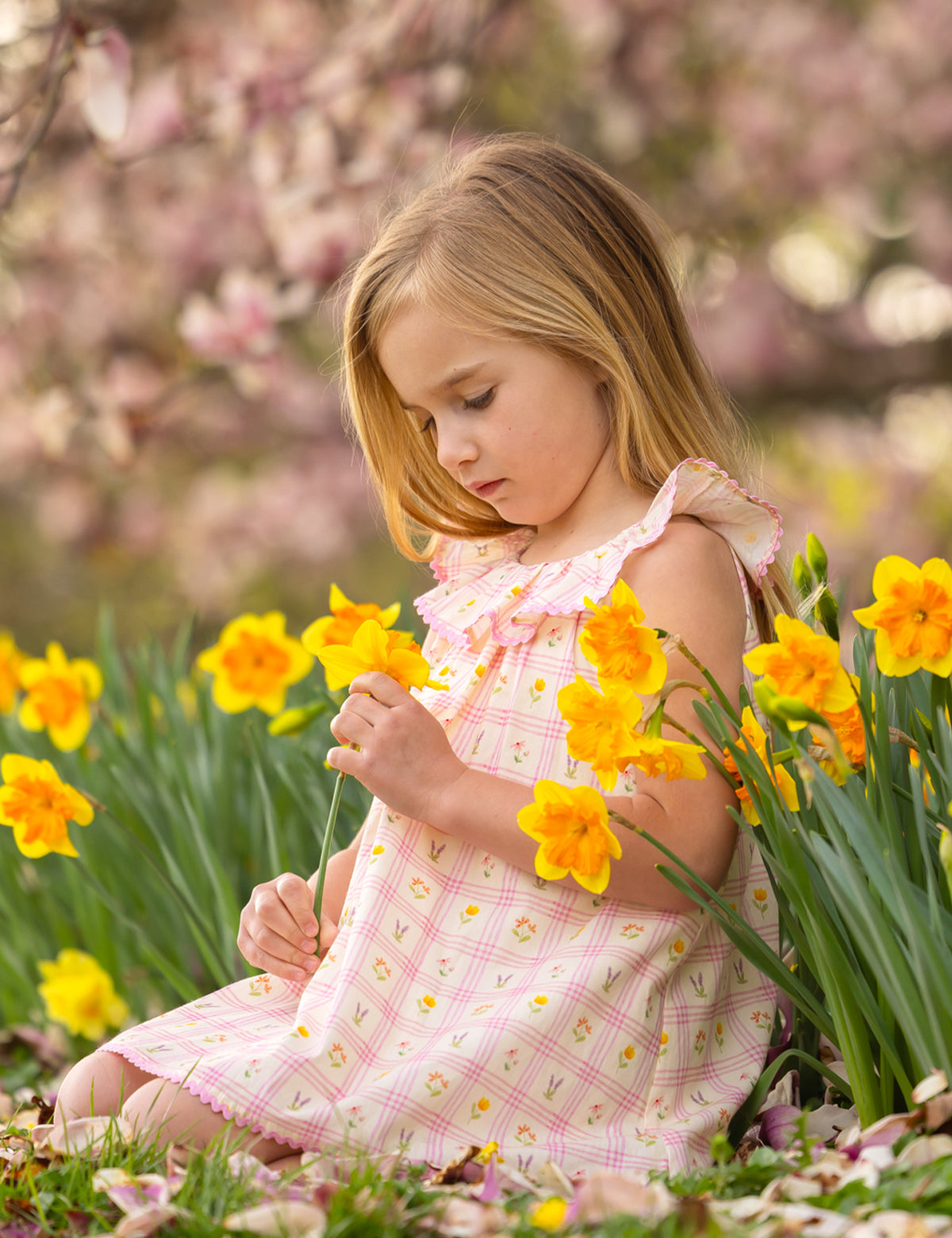 A young girl in the Mabel and Honey Ashley Cap Sleeve Dress sits in a garden among blooming yellow daffodils, gently holding a flower and looking down thoughtfully. Blossoming trees fill the background.