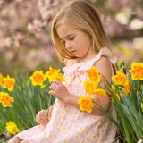 A young girl in the Mabel and Honey Ashley Cap Sleeve Dress sits in a garden among blooming yellow daffodils, gently holding a flower and looking down thoughtfully. Blossoming trees fill the background.