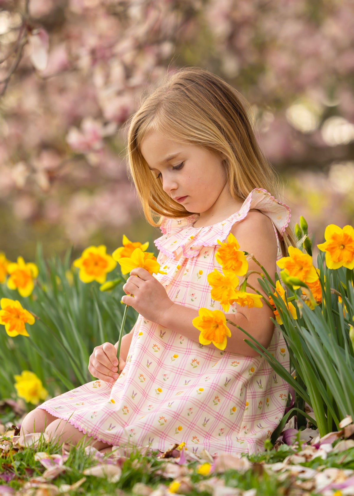 A young girl in the Mabel and Honey Ashley Cap Sleeve Dress sits in a garden among blooming yellow daffodils, gently holding a flower and looking down thoughtfully. Blossoming trees fill the background.
