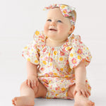 A smiling baby sits barefoot on a white surface, wearing the Mabel and Honey Petal Party Short Bubble Sleeve Romper with floral prints, ruffle details, and a matching headband, against a plain white background.