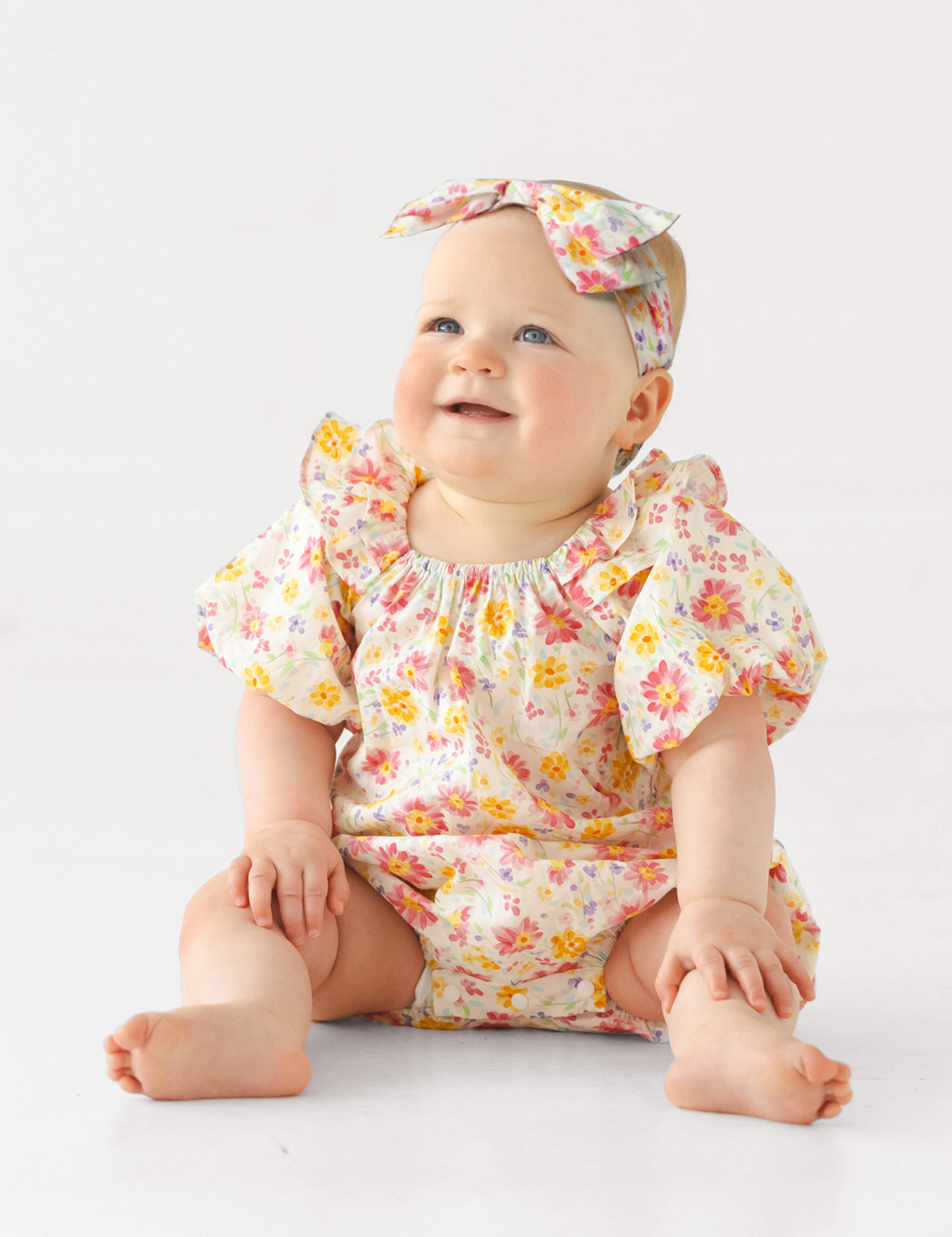 A smiling baby sits barefoot on a white surface, wearing the Mabel and Honey Petal Party Short Bubble Sleeve Romper with floral prints, ruffle details, and a matching headband, against a plain white background.