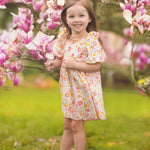 A young girl smiles among blooming pink magnolia flowers on a grassy lawn, wearing the Petal Party Short Bubble Sleeve Dress by Mabel and Honey, surrounded by soft sunlight and vibrant blossoms.