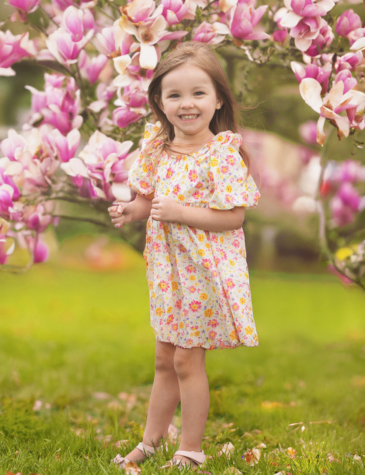 A young girl smiles among blooming pink magnolia flowers on a grassy lawn, wearing the Petal Party Short Bubble Sleeve Dress by Mabel and Honey, surrounded by soft sunlight and vibrant blossoms.