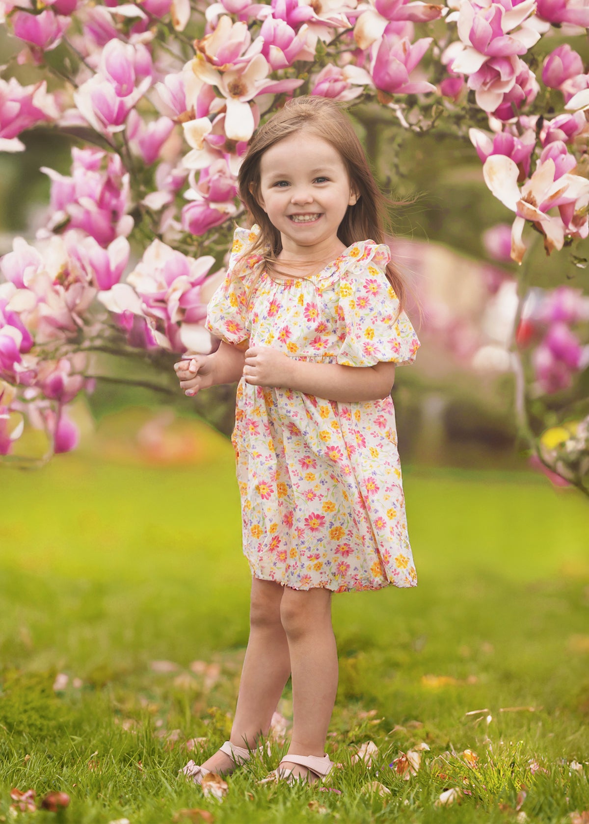A young girl smiles among blooming pink magnolia flowers on a grassy lawn, wearing the Petal Party Short Bubble Sleeve Dress by Mabel and Honey, surrounded by soft sunlight and vibrant blossoms.