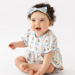 A smiling baby with curly dark hair sits barefoot on a white background, wearing the Mabel and Honey Blooms and Bows Short Gathered Ruffle Sleeve Romper with a matching bow headband.
