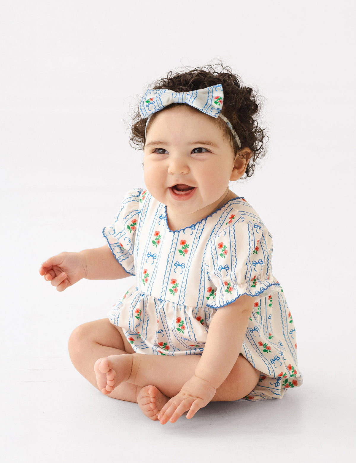 A smiling baby with curly dark hair sits barefoot on a white background, wearing the Mabel and Honey Blooms and Bows Short Gathered Ruffle Sleeve Romper with a matching bow headband.