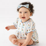 A smiling baby with curly dark hair sits barefoot on a white background, wearing the Mabel and Honey Blooms and Bows Short Gathered Ruffle Sleeve Romper with a matching bow headband.
