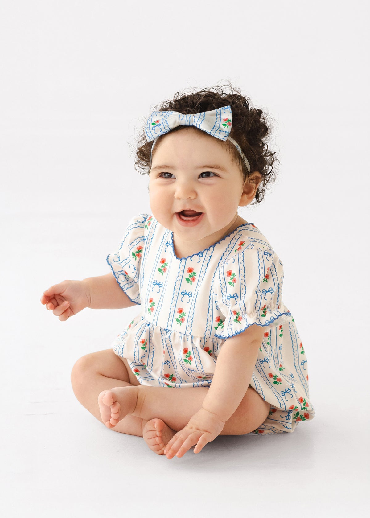 A smiling baby with curly dark hair sits barefoot on a white background, wearing the Mabel and Honey Blooms and Bows Short Gathered Ruffle Sleeve Romper with a matching bow headband.