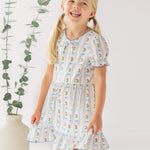 A young girl with blonde pigtails smiles indoors next to a tall vase of eucalyptus, wearing the Mabel and Honey Blooms and Bows Short Gathered Ruffle Sleeve Dress—lightweight, floral, and perfect for everyday wear.
