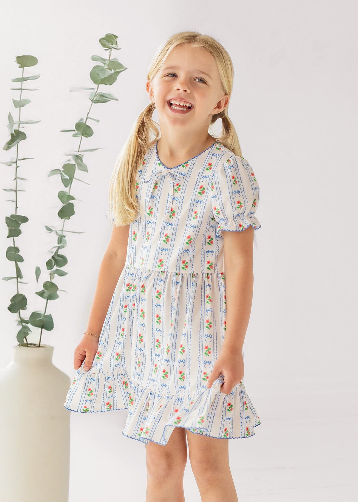 A young girl with blonde pigtails smiles indoors next to a tall vase of eucalyptus, wearing the Mabel and Honey Blooms and Bows Short Gathered Ruffle Sleeve Dress—lightweight, floral, and perfect for everyday wear.