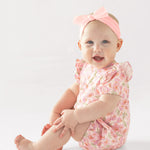 A smiling baby wears the Mabel and Honey Wild Savanna Short Gathered Sleeve Romper, a breathable pink floral outfit, with a matching bow headband, sitting on a white background and holding her leg.