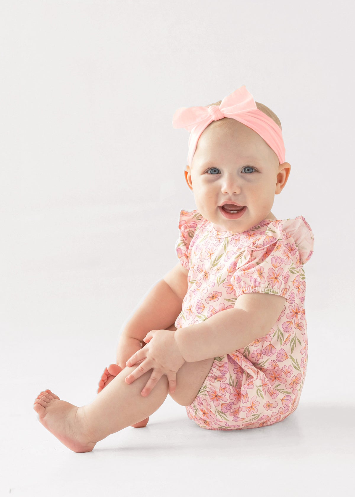 A smiling baby wears the Mabel and Honey Wild Savanna Short Gathered Sleeve Romper, a breathable pink floral outfit, with a matching bow headband, sitting on a white background and holding her leg.