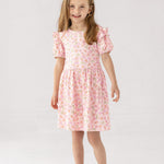 A smiling young girl with light brown hair stands against a white background, wearing the Mabel and Honey Wild Savanna Short Gathered Sleeve Dress and light pink sandals—a lightweight, floral outfit ideal for sunny days.