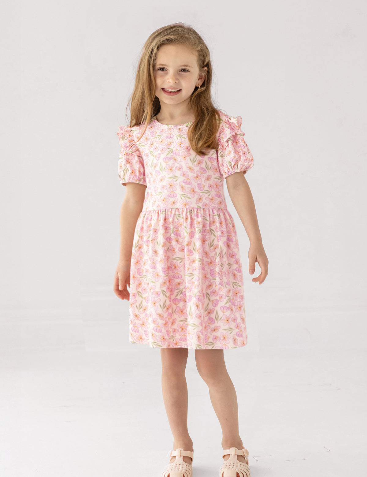A smiling young girl with light brown hair stands against a white background, wearing the Mabel and Honey Wild Savanna Short Gathered Sleeve Dress and light pink sandals—a lightweight, floral outfit ideal for sunny days.