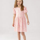 A smiling young girl with light brown hair stands against a white background, wearing the Mabel and Honey Wild Savanna Short Gathered Sleeve Dress and light pink sandals—a lightweight, floral outfit ideal for sunny days.