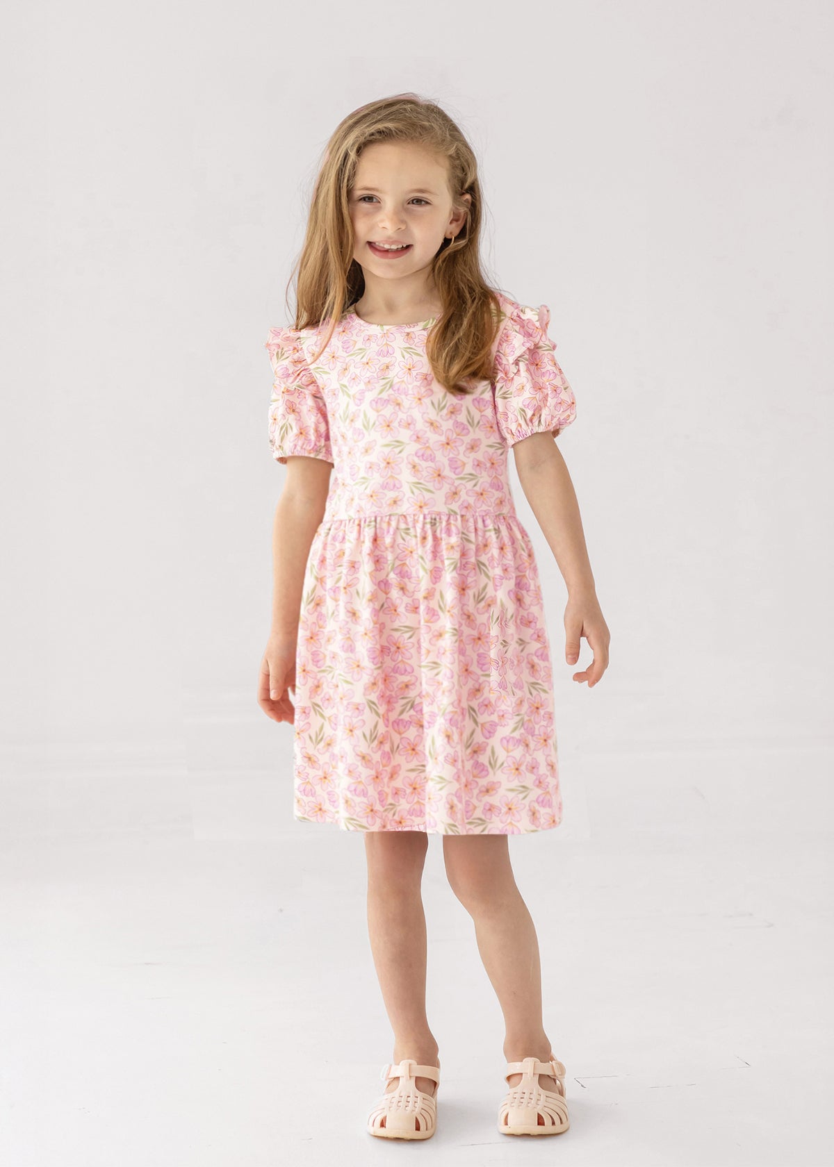 A smiling young girl with light brown hair stands against a white background, wearing the Mabel and Honey Wild Savanna Short Gathered Sleeve Dress and light pink sandals—a lightweight, floral outfit ideal for sunny days.