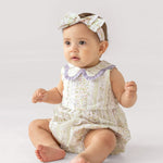 A baby wears the Graceful Garden Sleeveless Romper by Mabel and Honey, featuring floral prints and a purple ruffled collar, paired with a matching headband, while sitting on a white surface with a curious expression.