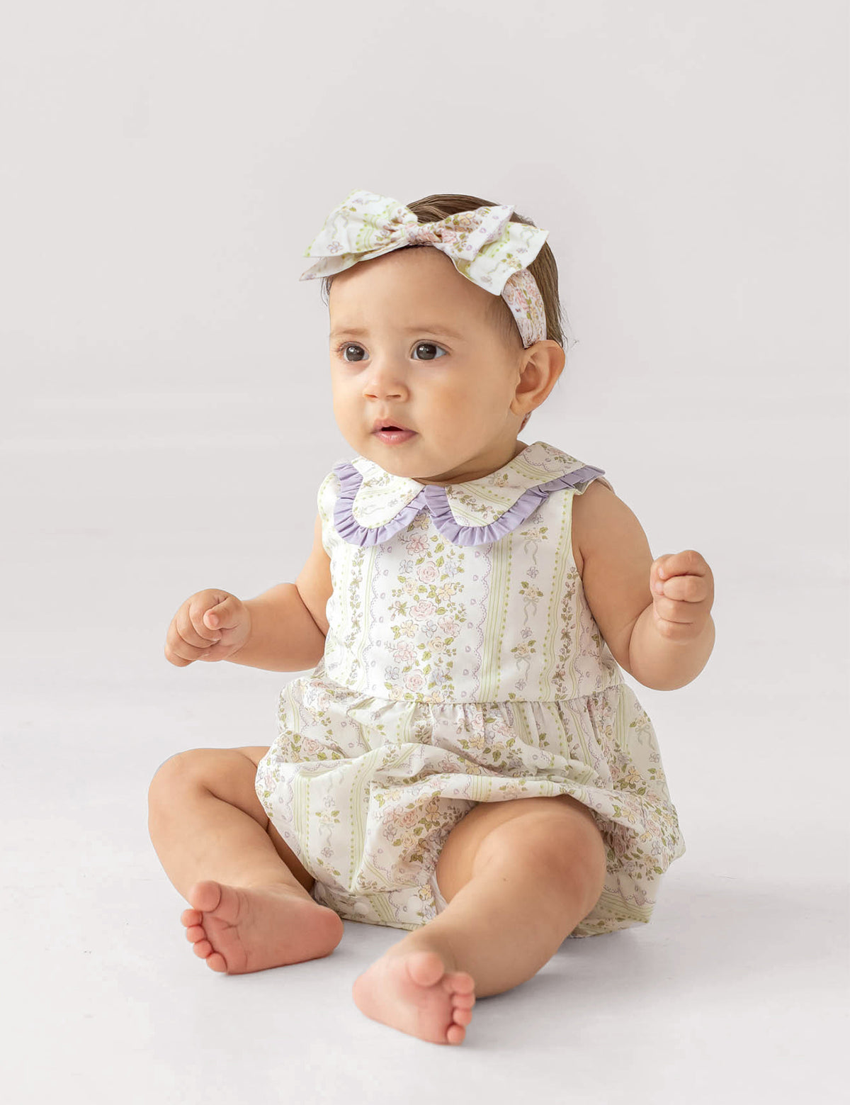 A baby wears the Graceful Garden Sleeveless Romper by Mabel and Honey, featuring floral prints and a purple ruffled collar, paired with a matching headband, while sitting on a white surface with a curious expression.