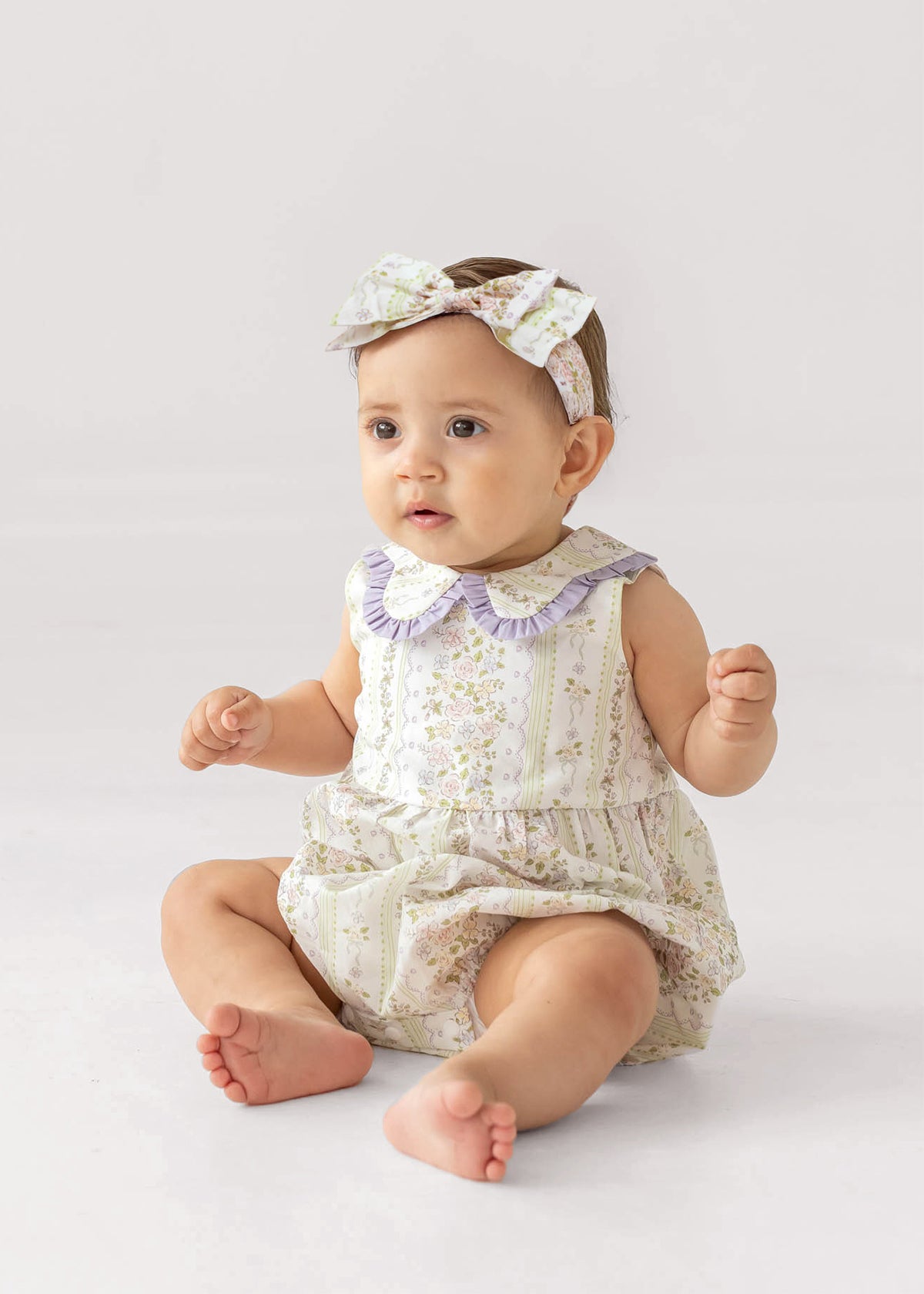 A baby wears the Graceful Garden Sleeveless Romper by Mabel and Honey, featuring floral prints and a purple ruffled collar, paired with a matching headband, while sitting on a white surface with a curious expression.