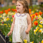 A young girl with long brown hair smiles in a sunny garden of tulips and daffodils, wearing the Graceful Garden Floral Dress by Mabel and Honey.