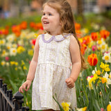 A young girl with long brown hair smiles in a sunny garden of tulips and daffodils, wearing the Graceful Garden Floral Dress by Mabel and Honey.