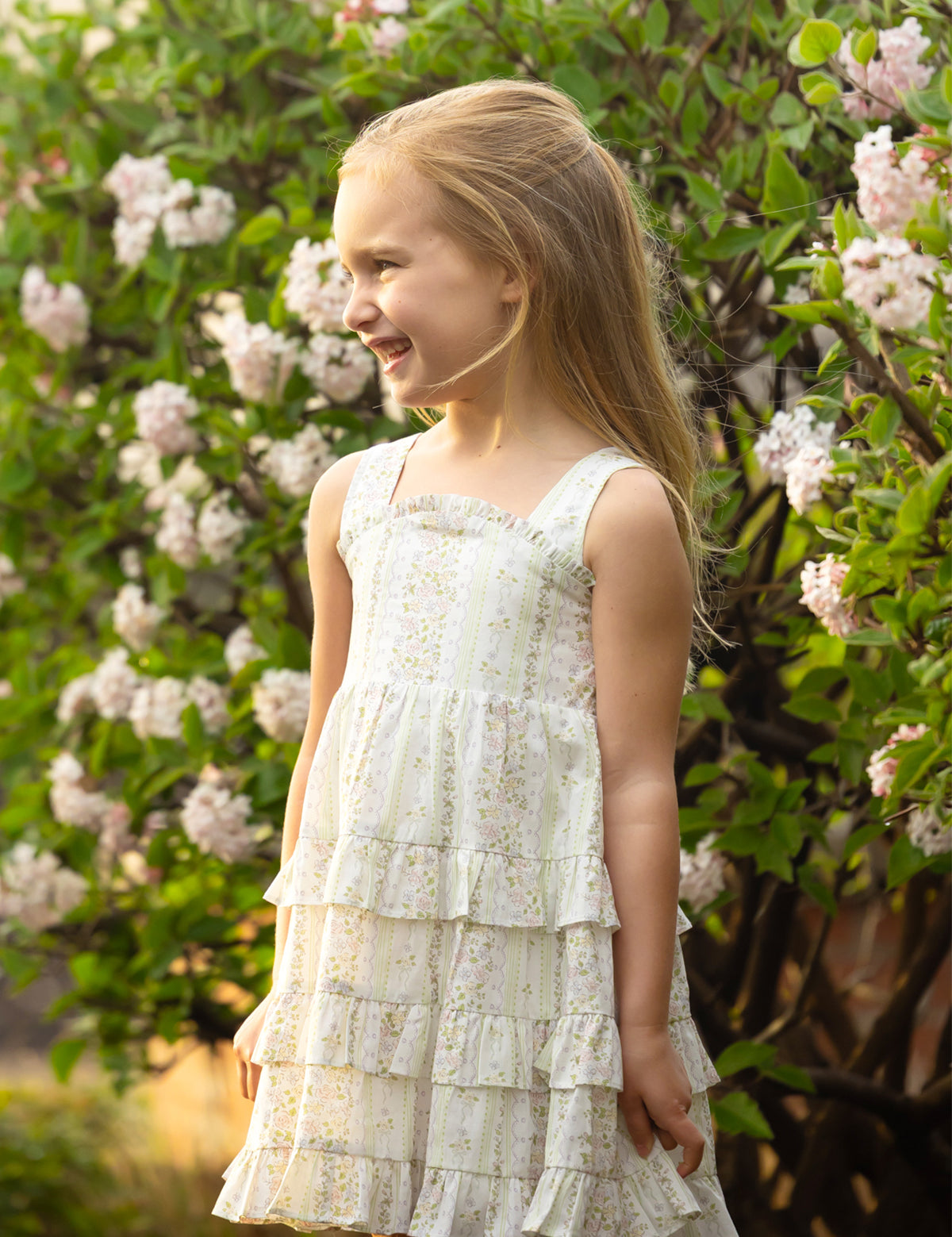 A young girl smiles outdoors in the Mabel and Honey Graceful Garden Sleeveless Floral Dress, surrounded by blooming white and pink flowers.