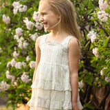 A young girl smiles outdoors in the Mabel and Honey Graceful Garden Sleeveless Floral Dress, surrounded by blooming white and pink flowers.