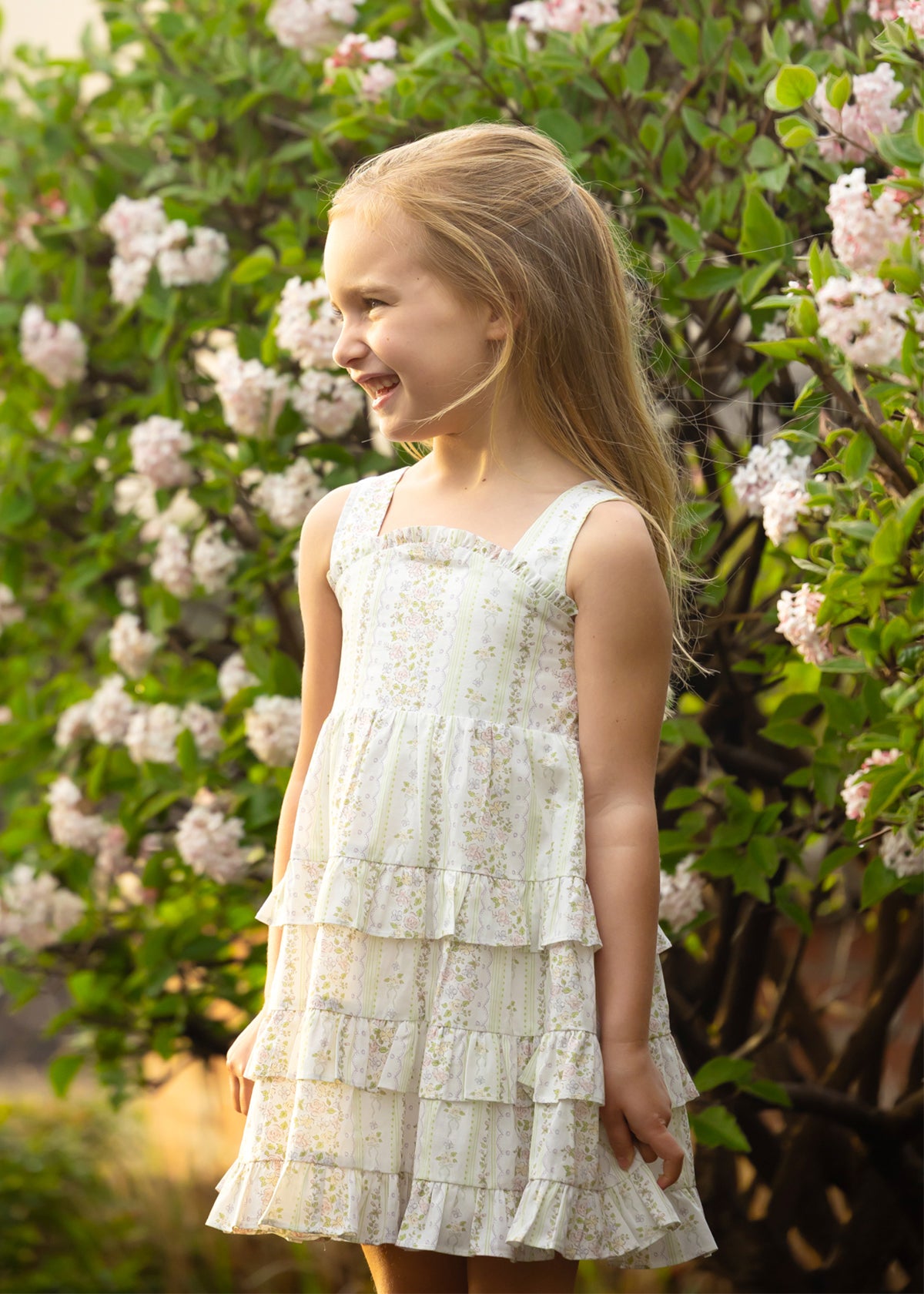 A young girl smiles outdoors in the Mabel and Honey Graceful Garden Sleeveless Floral Dress, surrounded by blooming white and pink flowers.