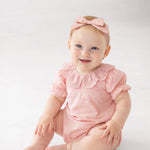 A baby wearing the Mabel and Honey Lacy Bowkisses Short Sleeve Romper with puffed sleeves, lace collar, and a matching pink bow headband sits on a white floor, smiling and looking to the side.
