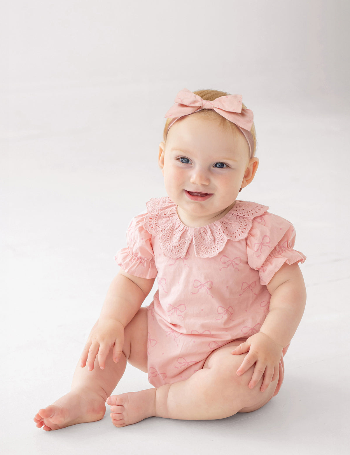 A baby wearing the Mabel and Honey Lacy Bowkisses Short Sleeve Romper with puffed sleeves, lace collar, and a matching pink bow headband sits on a white floor, smiling and looking to the side.