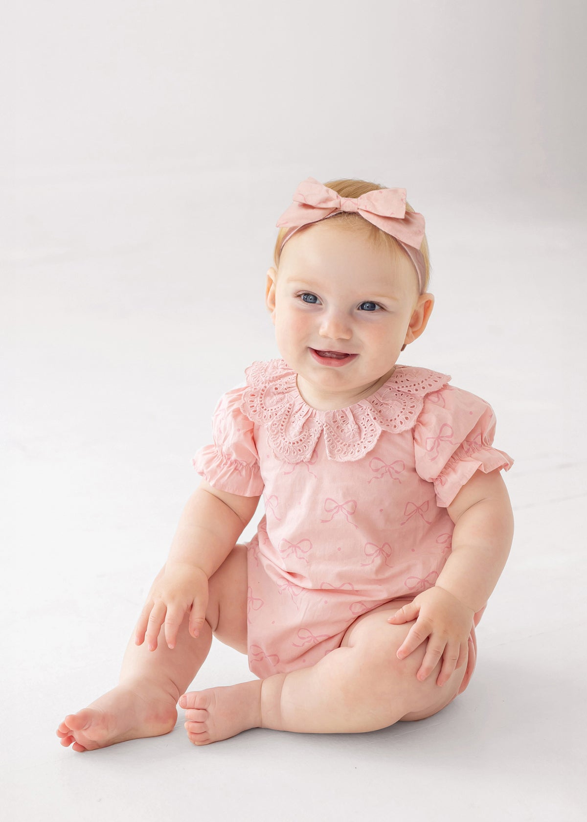 A baby wearing the Mabel and Honey Lacy Bowkisses Short Sleeve Romper with puffed sleeves, lace collar, and a matching pink bow headband sits on a white floor, smiling and looking to the side.