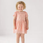 A young child with curly blond hair smiles in the Mabel and Honey Lacy Bowkisses Short Sleeve Dress, featuring puff sleeves and lace trim, paired with white sandals against a plain light background.