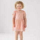 A young child with curly blond hair smiles in the Mabel and Honey Lacy Bowkisses Short Sleeve Dress, featuring puff sleeves and lace trim, paired with white sandals against a plain light background.