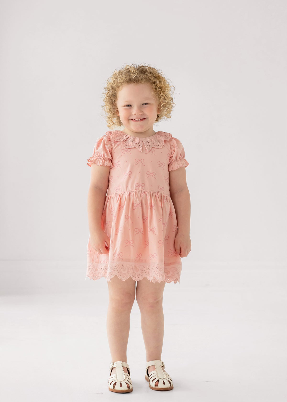 A young child with curly blond hair smiles in the Mabel and Honey Lacy Bowkisses Short Sleeve Dress, featuring puff sleeves and lace trim, paired with white sandals against a plain light background.