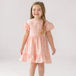 A young girl with long light brown hair smiles in the Mabel and Honey Lacy Bowkisses Puff Sleeve Dress, featuring ruffled layers, paired with white sandals against a plain white background.