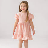 A young girl with long light brown hair smiles in the Mabel and Honey Lacy Bowkisses Puff Sleeve Dress, featuring ruffled layers, paired with white sandals against a plain white background.