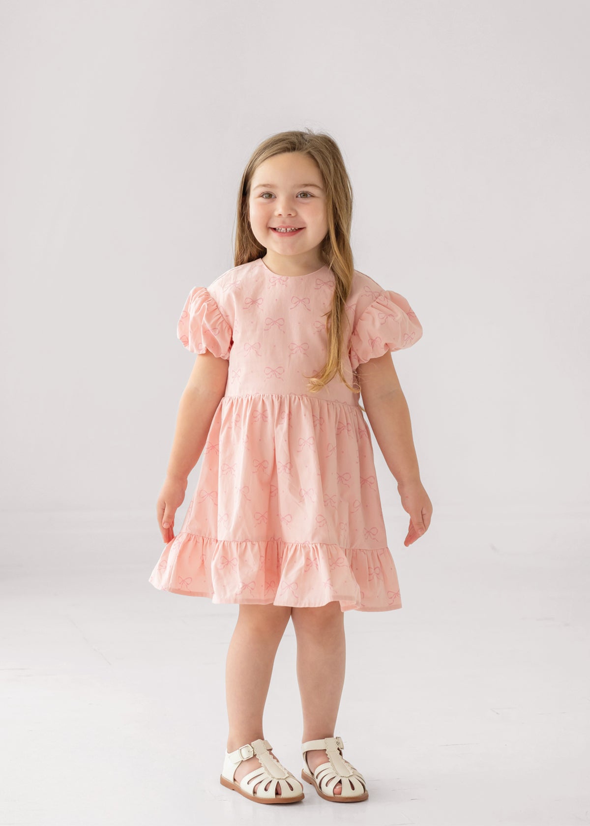 A young girl with long light brown hair smiles in the Mabel and Honey Lacy Bowkisses Puff Sleeve Dress, featuring ruffled layers, paired with white sandals against a plain white background.